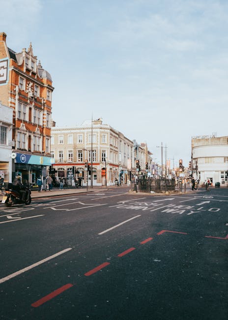 A wide urban street scene in daylight showing a junction with a mix of historic and modern buildings on the left and right sides, featuring multi-storey brick and stucco facades with decorative architectural details. The street has clearly marked pedestrian crossings, with a few pedestrians walking along the sidewalks and waiting at the crossings. In the foreground, there is an empty section of the road with painted lines and traffic markings, including red dashed lines indicating a bus stop or restricted zone. To the left, a parked scooter is visible on the pavement near a low curb. The background extends into a busy commercial area with shopfronts, signs, and streetlights, under a partly cloudy sky. This scene is typical of city centre environments where private waste removal services, such as those offered by Waste Disposal Camden, may carry out on-site clearance of rubbish in an alternative to local authority collection, facilitating independent waste management solutions.