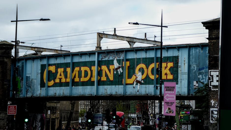 A street scene showing a row of historic brick buildings with large arched windows and traditional black lampposts along the pavement. One building displays a small orange sign for a juice shop. A vintage-style clock is mounted on a pole in front of the buildings. In the background, a blue railway bridge features yellow lettering reading 'Camden Lock,' with railway tracks and overhead electric wires above. Several pedestrians are walking along the sidewalk and crossing the street, some dressed in dark clothing. Parked cars are lined on the street, and a tall streetlamp and trees with budding leaves are visible to the right, indicating a spring setting. The environment suggests an urban area with a mix of commercial and transportation elements, typical of private or alternative waste collection contexts like rubbish removal services in Camden, such as Waste Disposal Camden, in a lively and active part of the town.