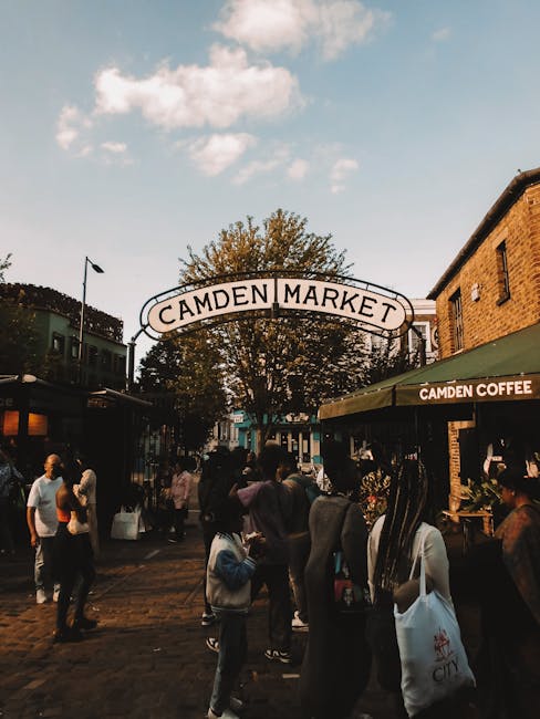 The image depicts the entrance to Camden Market on a busy day, with a large white archway sign reading 'CAMDEN MARKET' positioned centrally above the pathway. In the foreground, numerous pedestrians are walking along the paved area, some carrying shopping bags, while others browse or converse. To the right, a brick building with a green awning labeled 'CAMDEN COFFEE' extends over outdoor seating, with a few people gathered nearby. On the left, additional shops with large glass windows can be seen, partially obscured by the crowd. The environment has a lively atmosphere, shaded partly by the branches of a large tree situated behind the arch, with a clear sky and a few clouds overhead contributing to daylight conditions. The scene evokes a typical outdoor shopping and socialising space, often serviced by private waste collection companies like Waste Disposal Camden, who handle rubbish removal from commercial premises in busy market areas to support cleanliness and tidy environment management during daily operations.