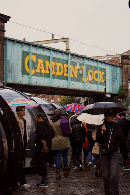 In the foreground, a group of people are gathered on a wet cobbled street, many holding umbrellas in various colors, including a prominent Union Jack design. The individuals are dressed in casual, weather-appropriate clothing, with some wearing jackets and hoodies, and appear to be waiting or walking along the area. To the left, there are several dark-colored, curved bus shelters or coverings, with some people standing nearby. Behind the crowd, there is a series of parked vehicles, including a car partly visible at the edge of the frame. Above the scene, an aged, weathered metal railway bridge painted in a light blue-turquoise hue spans across the street, bearing large, partially peeling yellow and orange lettering spelling “CANDEN LOCK.” The environment suggests an urban setting close to a market or busy transit area, with the weather contributing to a slightly damp and overcast atmosphere. The scene illustrates a typical busy street scene with people engaging in private disposal or on-site clearance activities, often associated with alternative waste handling services like waste disposal Camden, visible in the context of rubbish removal.
