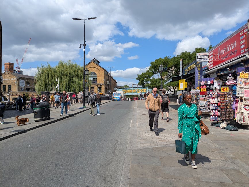 The image depicts the entrance to Camden Market on a busy day, with a large white archway sign reading 'CAMDEN MARKET' positioned centrally above the pathway. In the foreground, numerous pedestrians are walking along the paved area, some carrying shopping bags, while others browse or converse. To the right, a brick building with a green awning labeled 'CAMDEN COFFEE' extends over outdoor seating, with a few people gathered nearby. On the left, additional shops with large glass windows can be seen, partially obscured by the crowd. The environment has a lively atmosphere, shaded partly by the branches of a large tree situated behind the arch, with a clear sky and a few clouds overhead contributing to daylight conditions. The scene evokes a typical outdoor shopping and socialising space, often serviced by private waste collection companies like Waste Disposal Camden, who handle rubbish removal from commercial premises in busy market areas to support cleanliness and tidy environment management during daily operations.